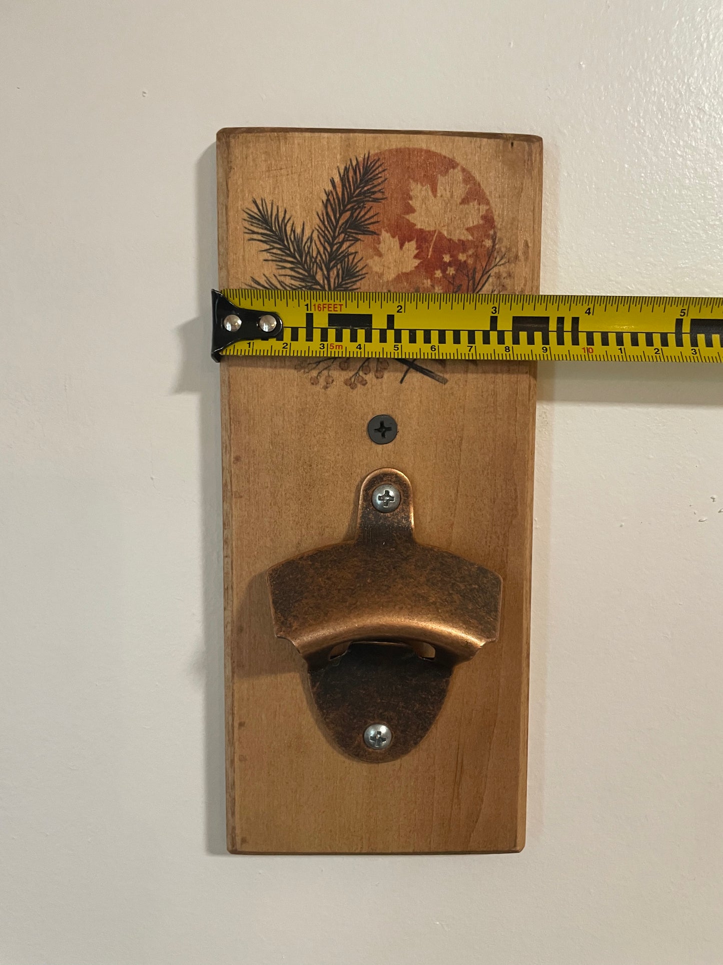 A rustic wooden wall-mounted bottle opener with a copper-toned metal opener attached to a light brown wood plaque featuring a printed autumn leaf and pine branch design. A yellow tape measure is stretched horizontally across the top of the plaque to show its width against a plain off-white wall.