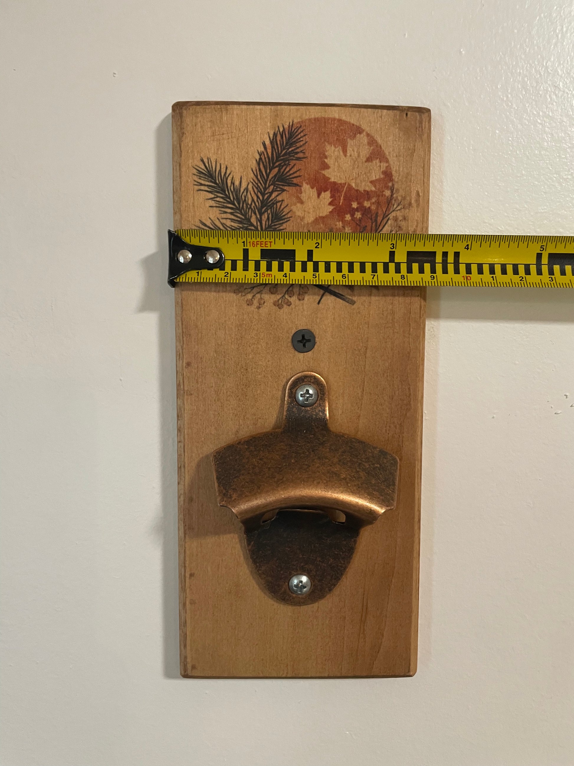 A rustic wooden wall-mounted bottle opener with a copper-toned metal opener attached to a light brown wood plaque featuring a printed autumn leaf and pine branch design. A yellow tape measure is stretched horizontally across the top of the plaque to show its width against a plain off-white wall.