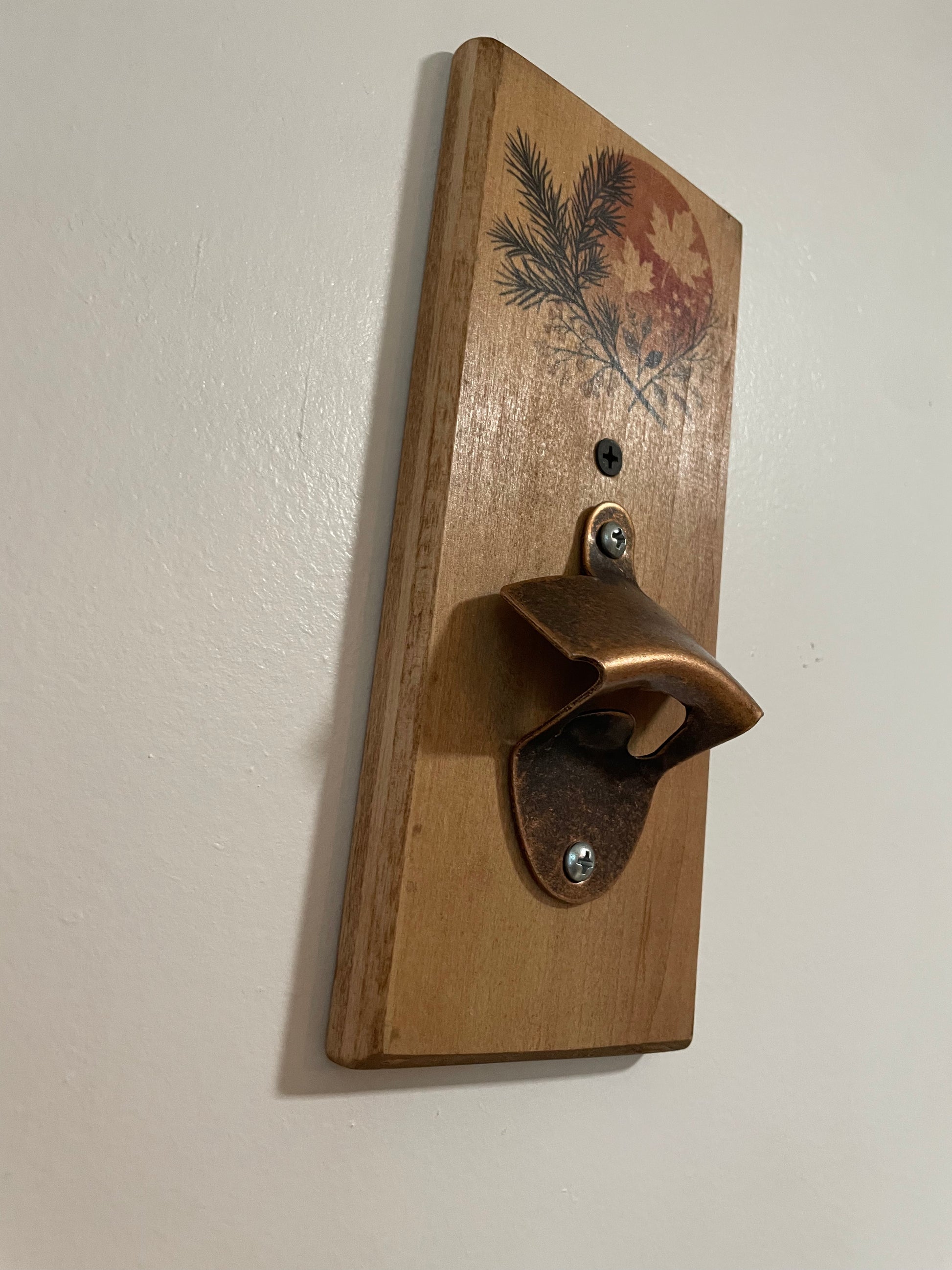 A rustic wall-mounted bottle opener made from a natural wood plank with a printed pine and maple leaf design, featuring a copper-colored metal opener attached with screws.