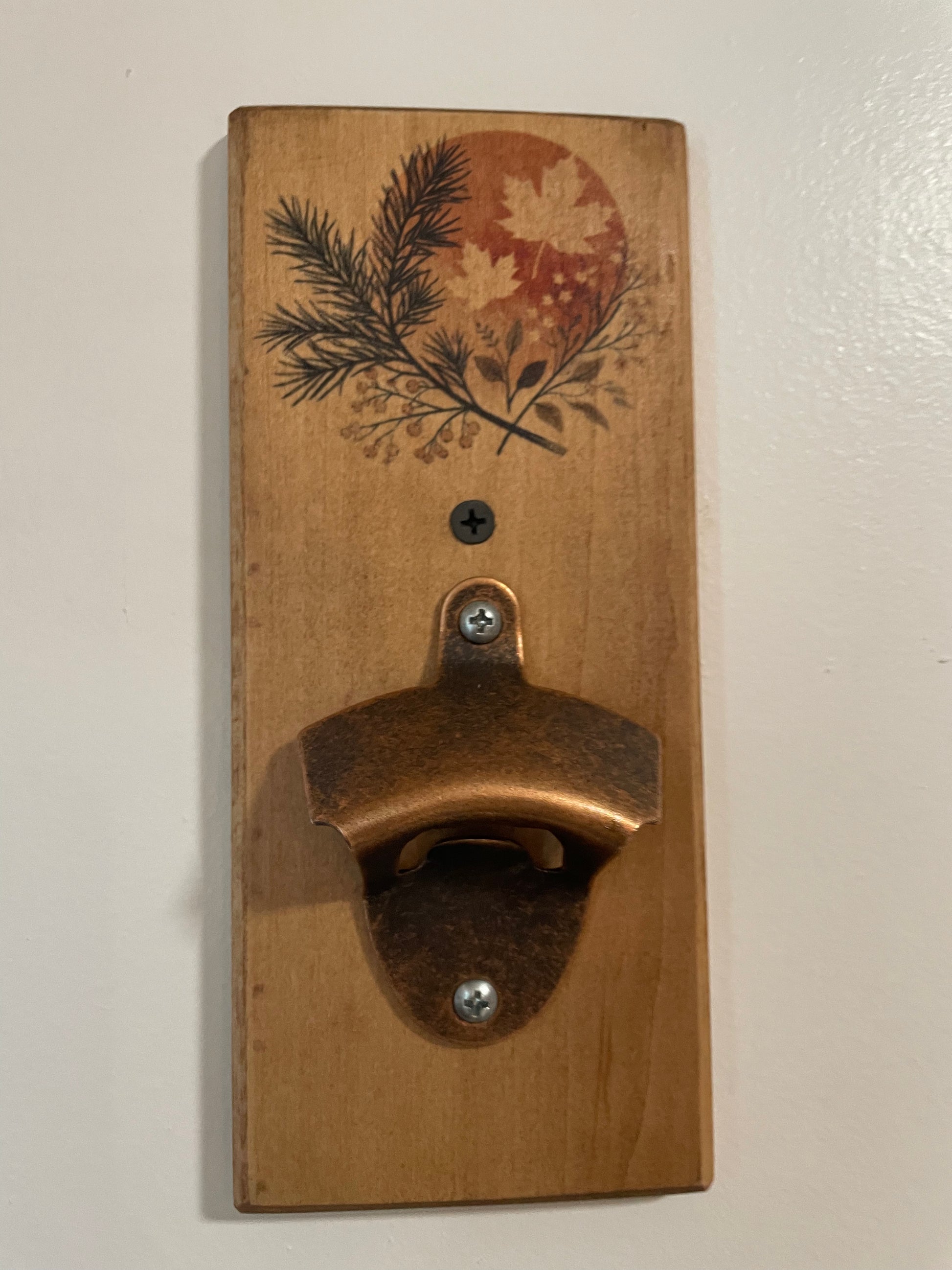 A rustic wall-mounted bottle opener made from a rectangular wooden board with a decorative pine branch and maple leaf design above a copper-toned metal opener, shown against an off-white wall.