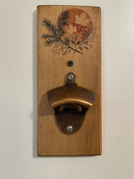 A rustic wall-mounted bottle opener made from a rectangular wooden plaque with a copper-toned metal opener attached. The wood features a printed botanical design of pine branches and maple leaves over a muted orange circle near the top.
